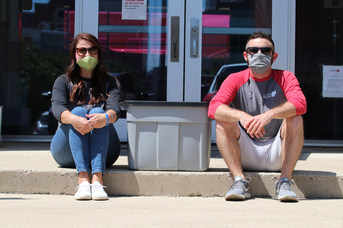 Two college staff members sit outside the PAES building with a plastic bin of food between them