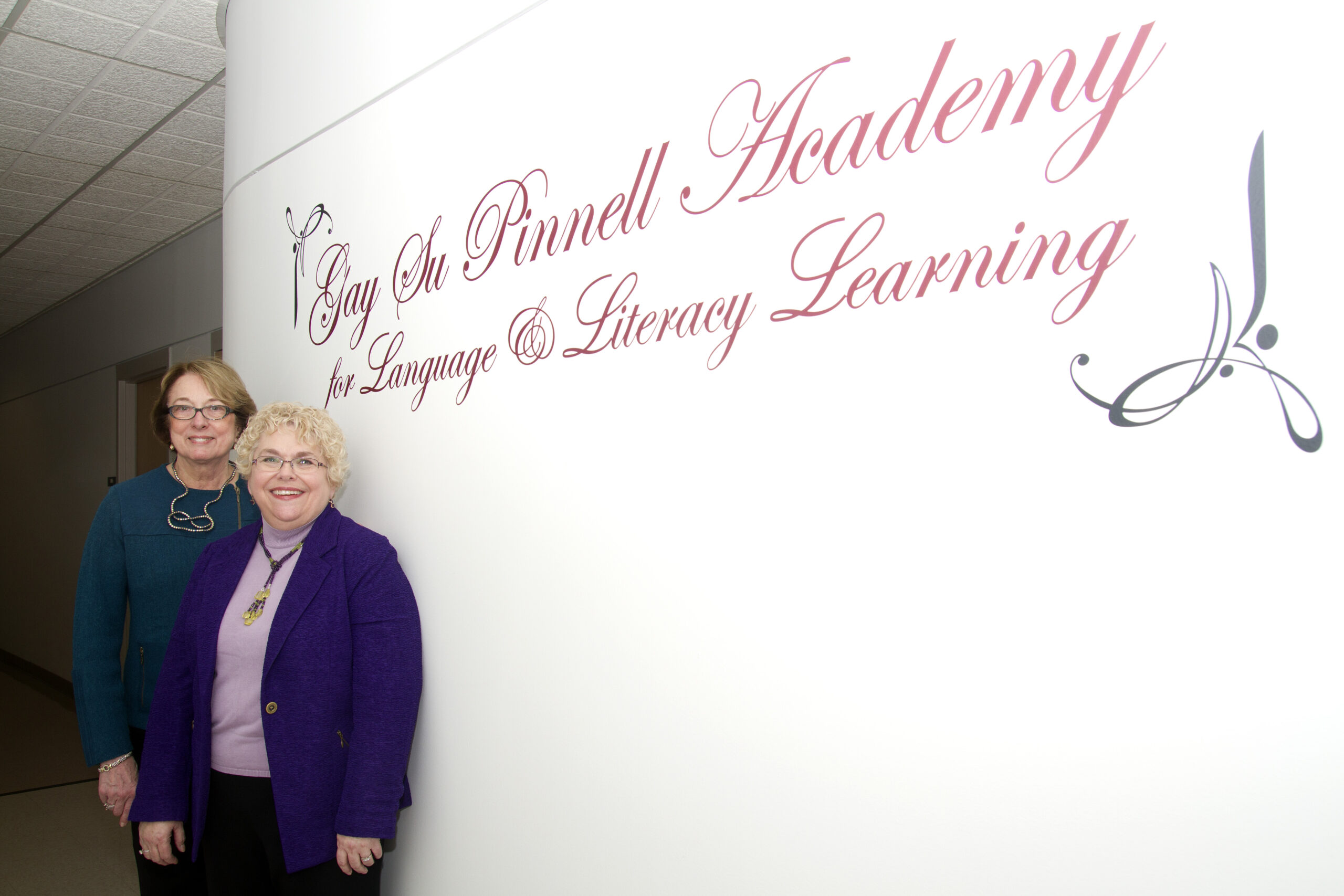 Two women stand near a wall with writing that says "Gay Su Pinnell Academy for Language and Literacy Learning"