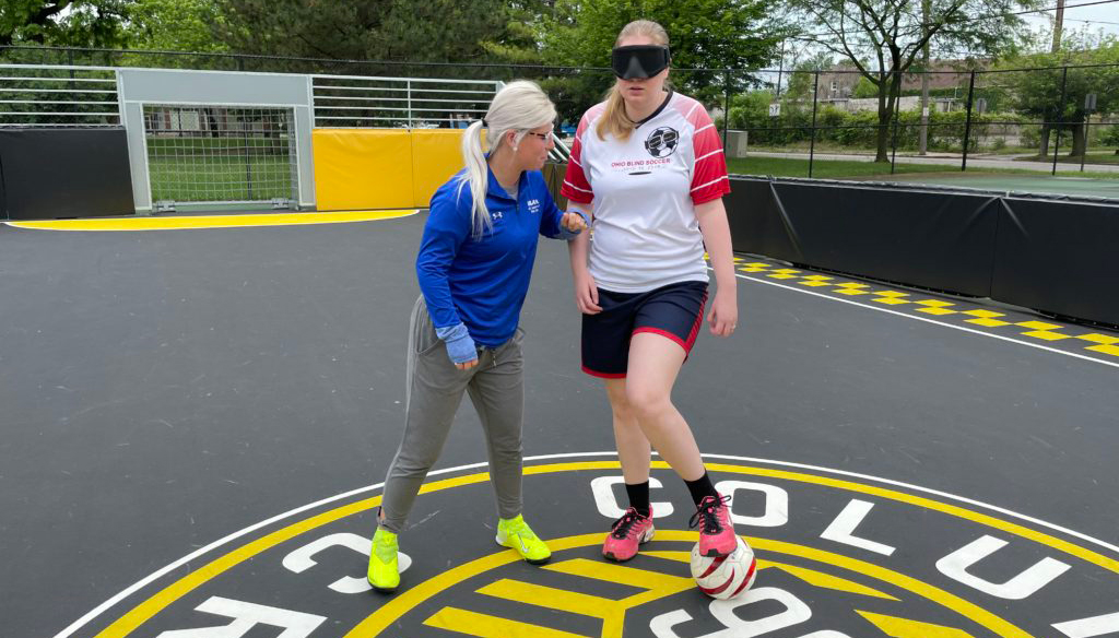 Two women practicing soccer on Columbus Crew mat, one wearing a blindfold