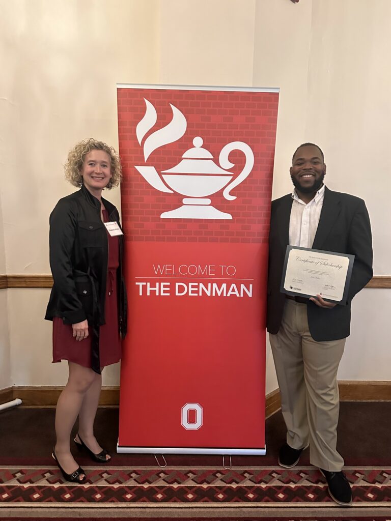 Elizabeth Arthur and John Fuller standing by a sign reading "Welcome to The Denman" award ceremony