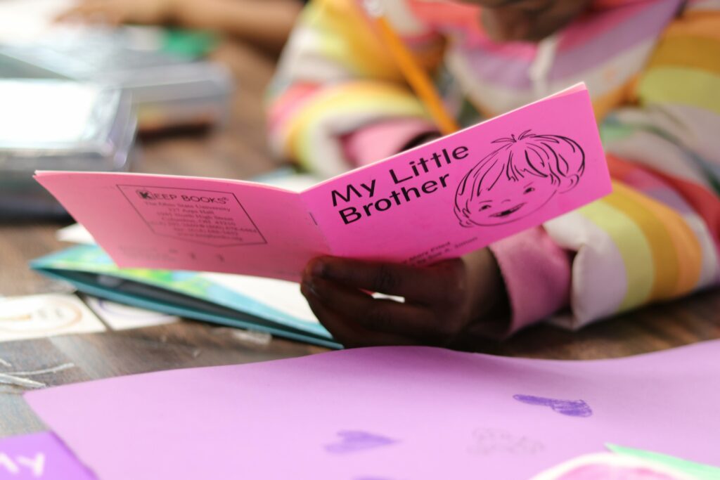Close up of child's hand holding a pink book with the title "My Little Brother" at a school desk
