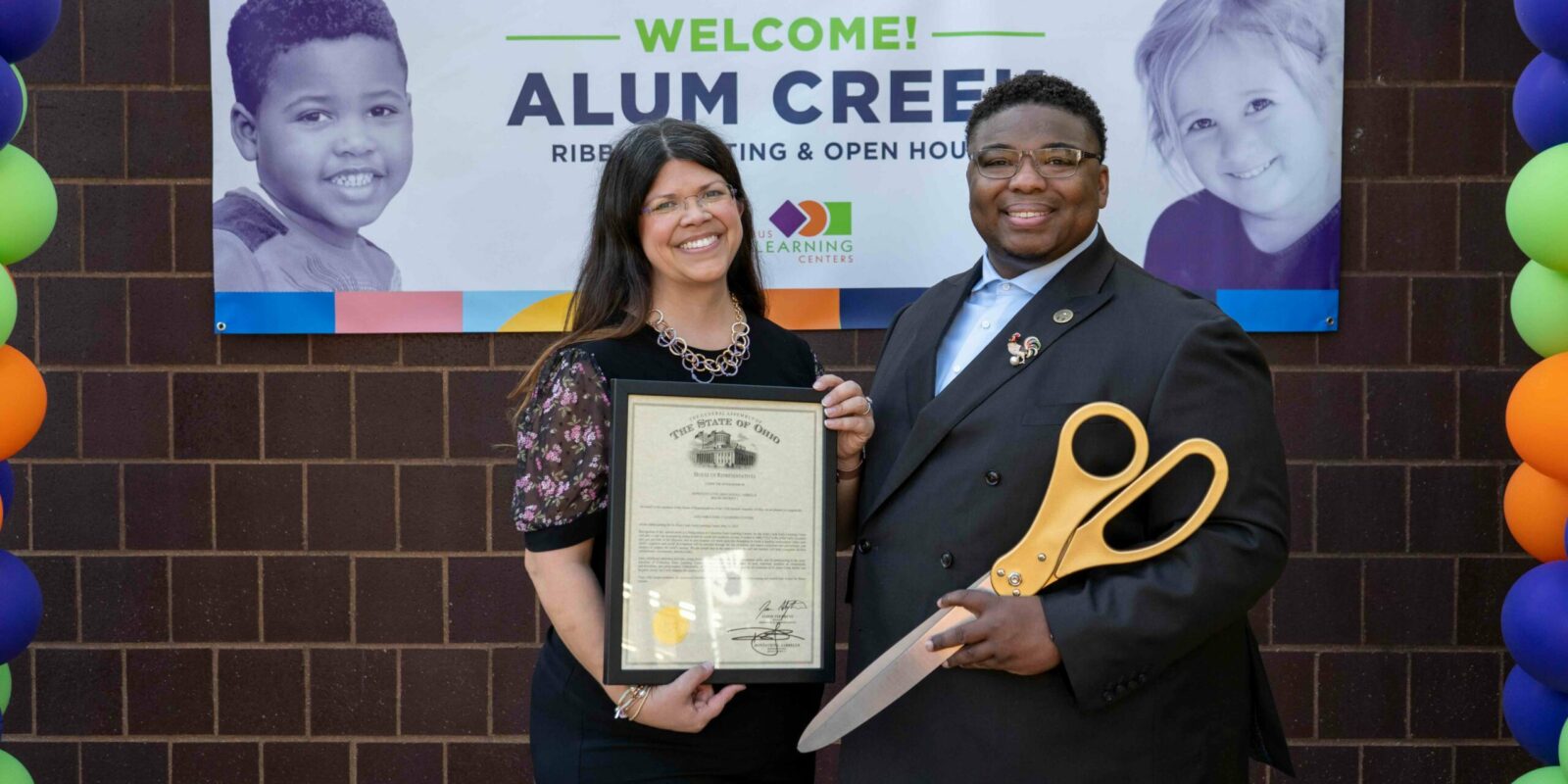 Gina Ginn holding an award at a ribbon cutting ceremony. Ohio Representative Dontavius Jarrells stands beside her with large scissors