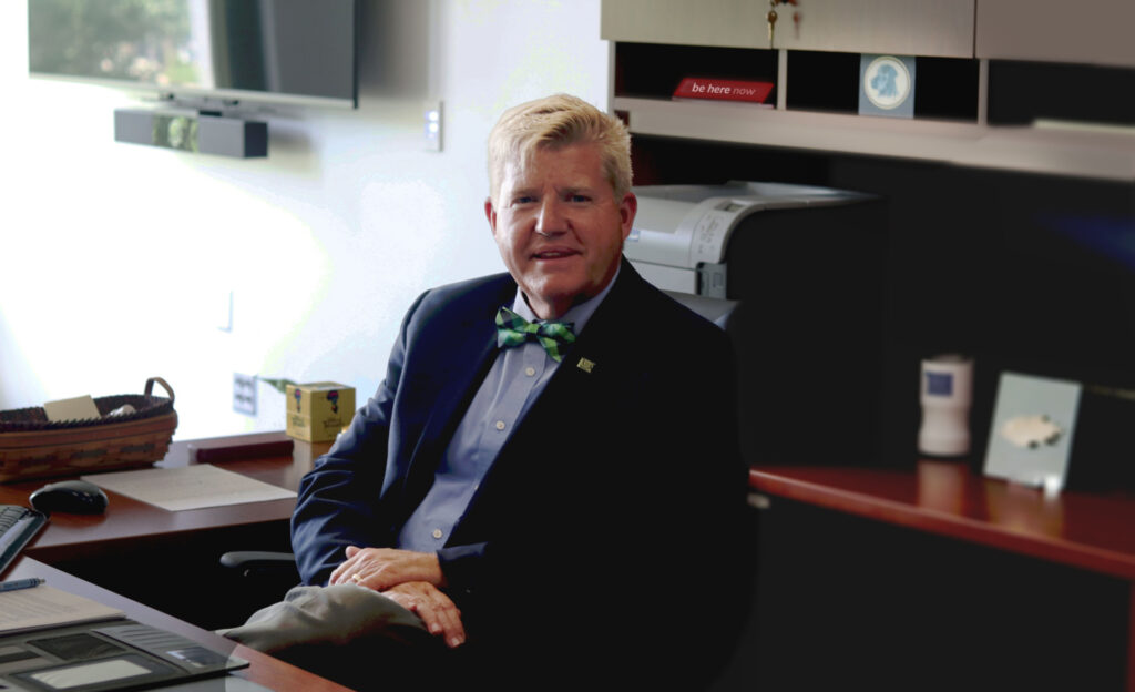 John Berry at his desk with a sign reading "be here now" in the background