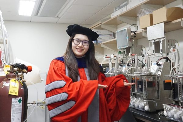 Shiqi Zhang in graduate regalia showing off the equipment in the lab.