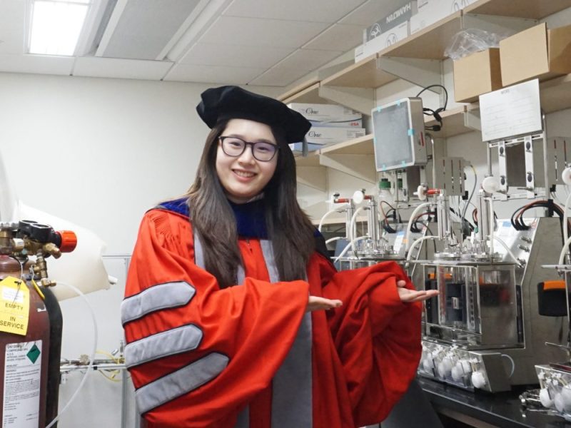 Shiqi Zhang in graduate regalia showing off the equipment in the lab.
